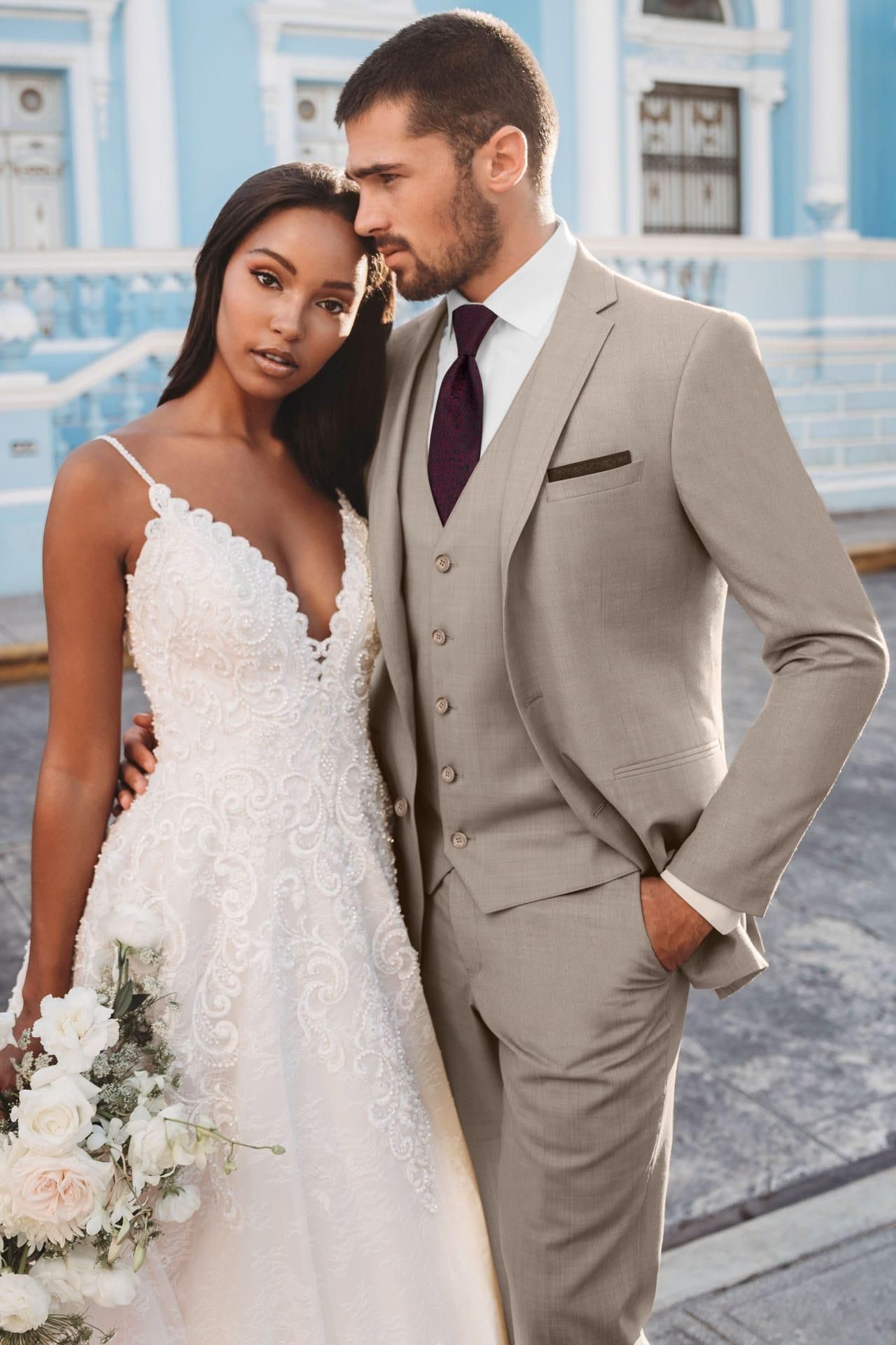 Bride and groom embrace outdoors; she wears a white gown, he wears a tan suit.