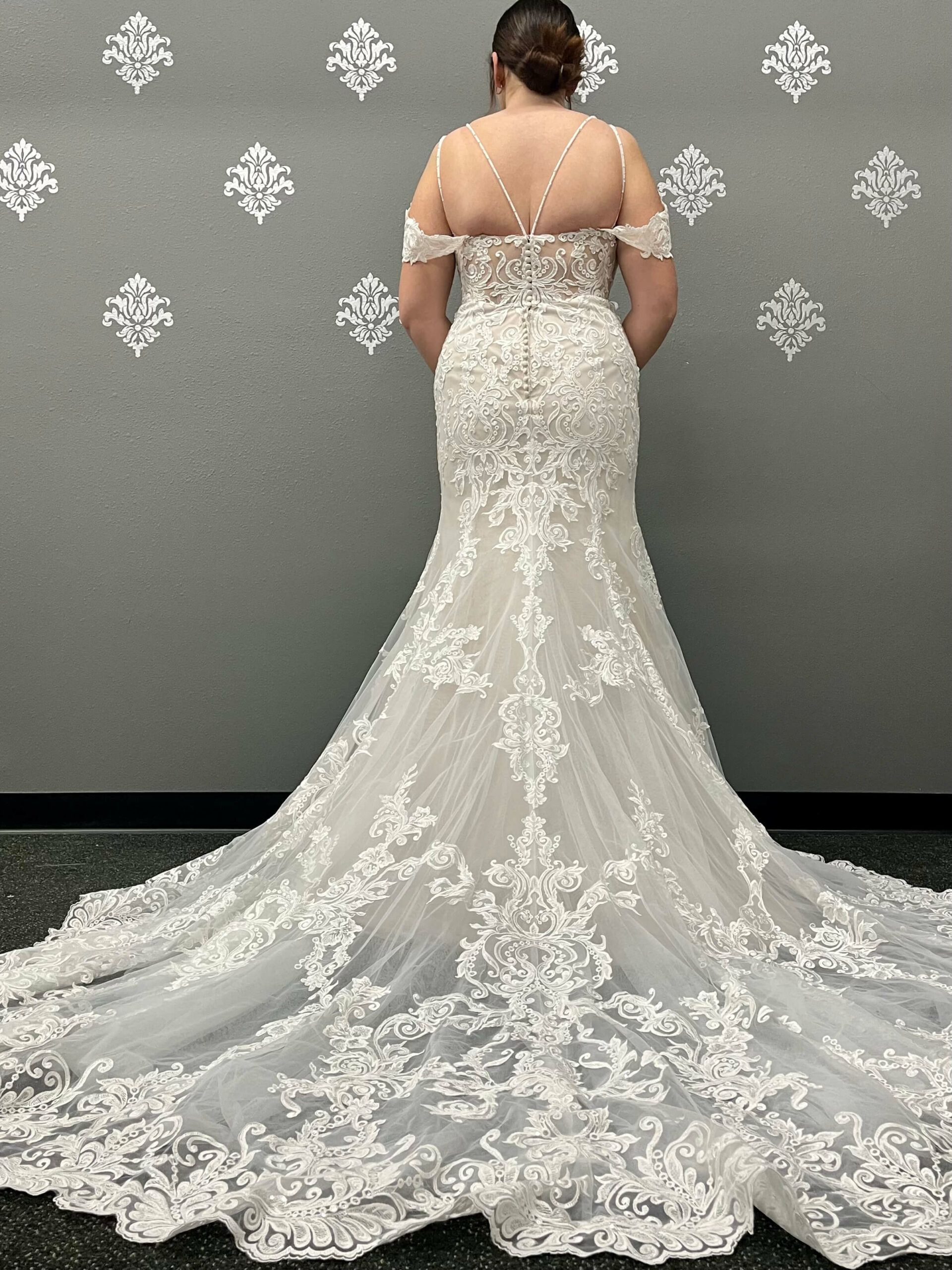 Bride in a white lace mermaid wedding dress, back view, standing in front of a gray wall with decorative patterns.