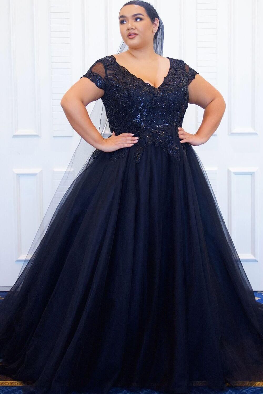 Woman in a black, sequined ball gown with a deep V-neck and tulle skirt, posing with hands on hips.
