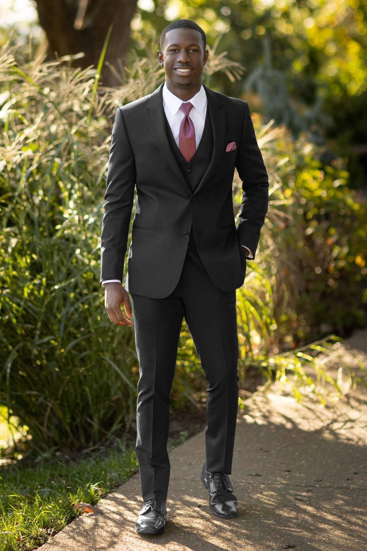 Man in a black suit smiles outdoors, next to greenery.