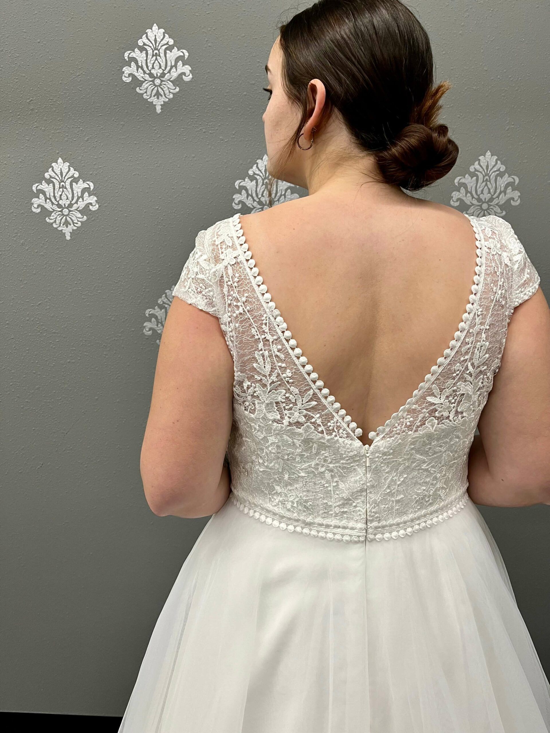 Woman in a white wedding dress, back view, with lace bodice and low back, standing in front of a gray wall.