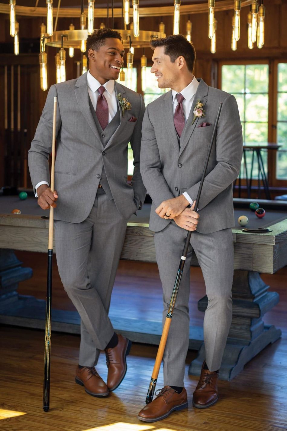 Two men in gray suits smile, holding pool cues near a pool table.