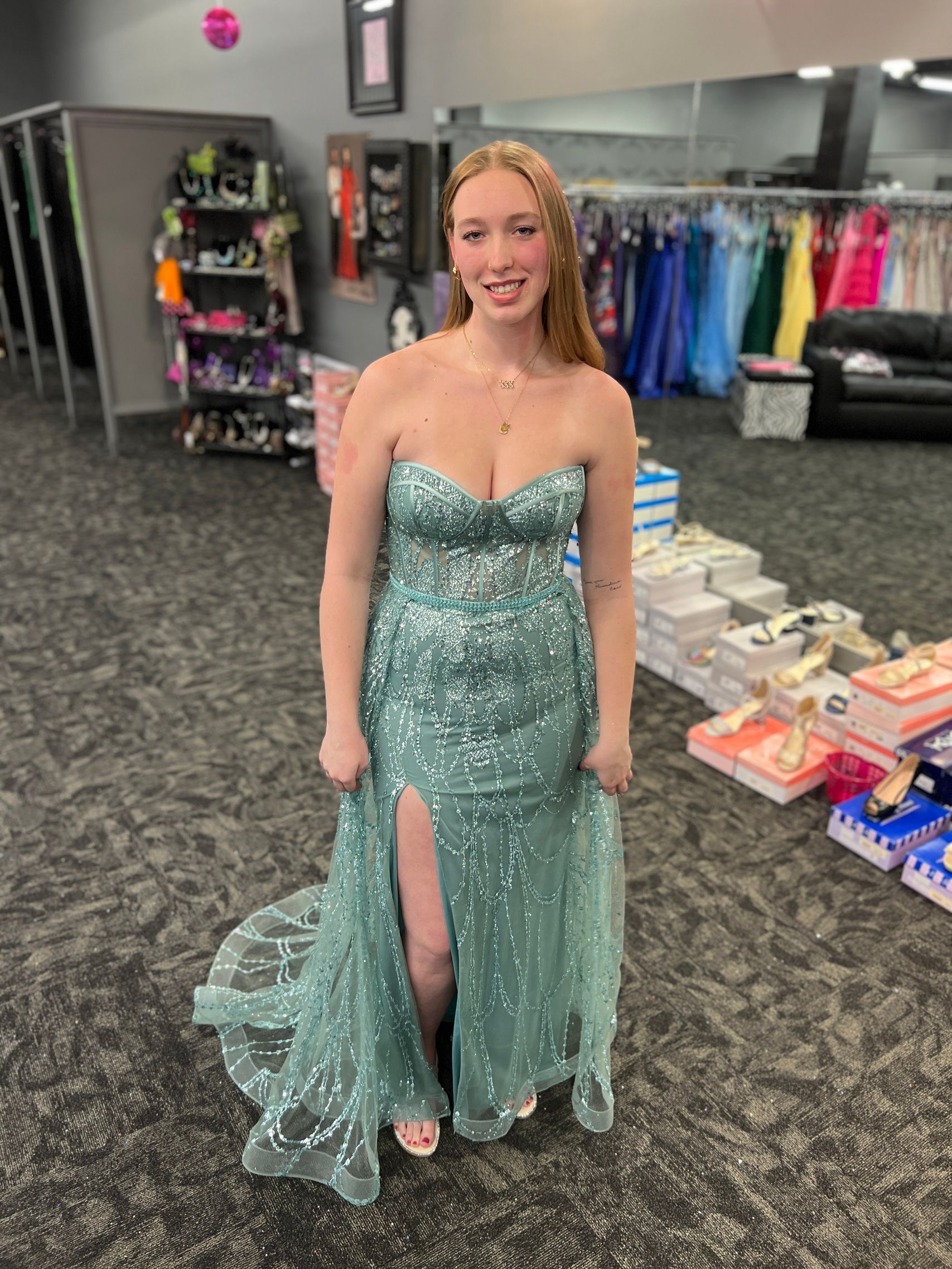 Woman in a teal strapless formal gown with a high slit, posing in a dress shop.
