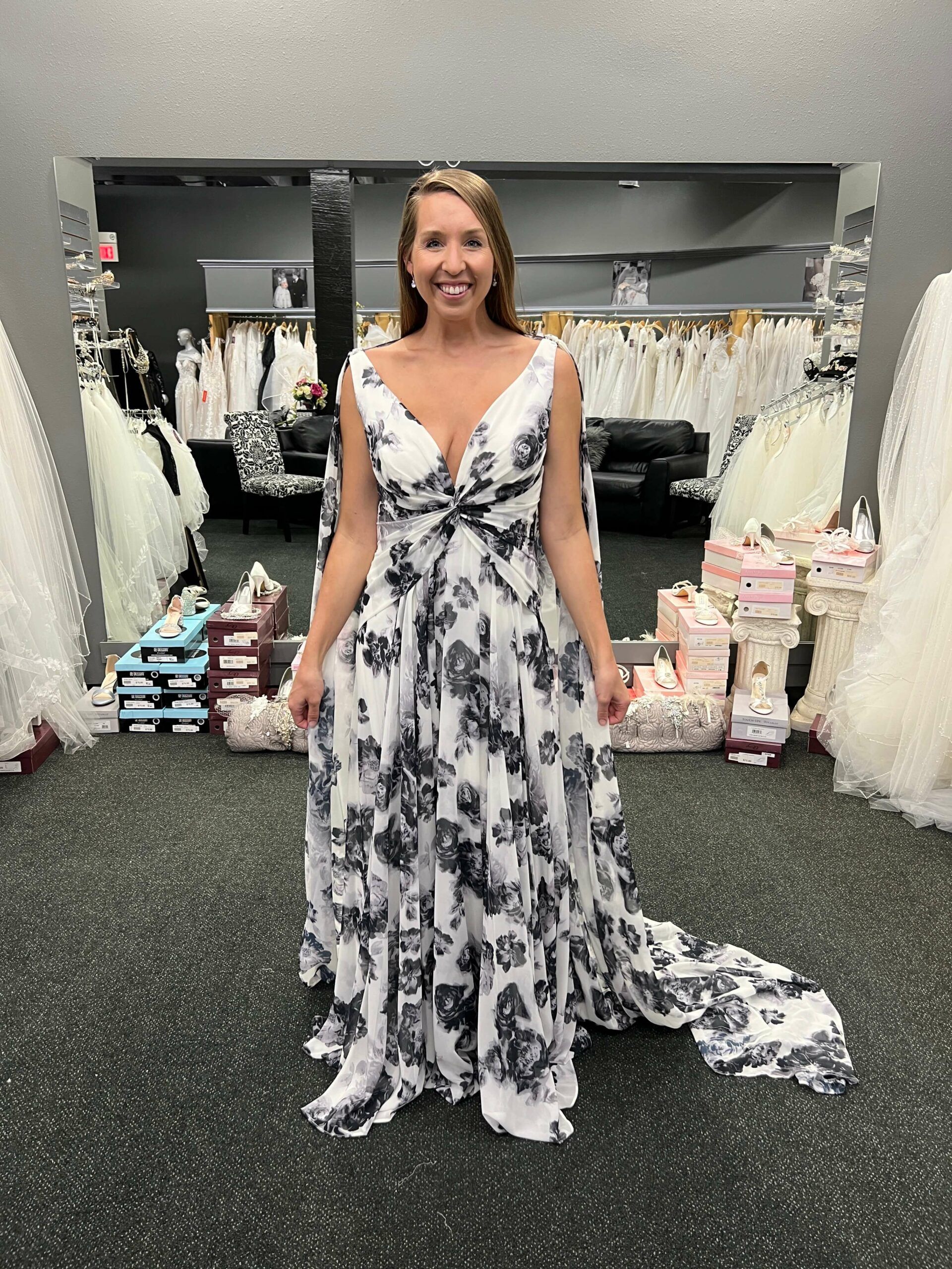 Woman in floral wedding dress smiles in a bridal shop, train trailing behind her.