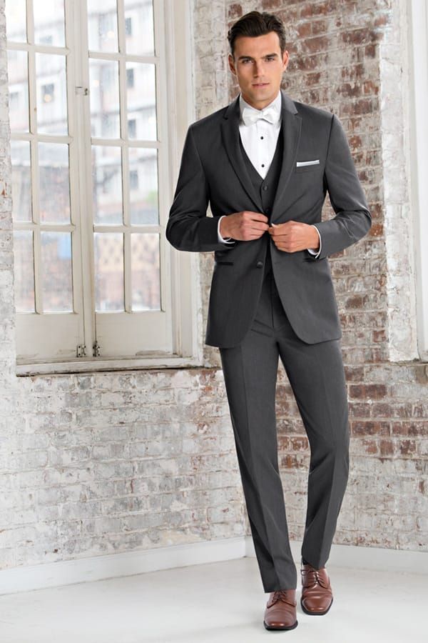 Man in a gray suit, white shirt, and bow tie stands by a window, buttoning his jacket.