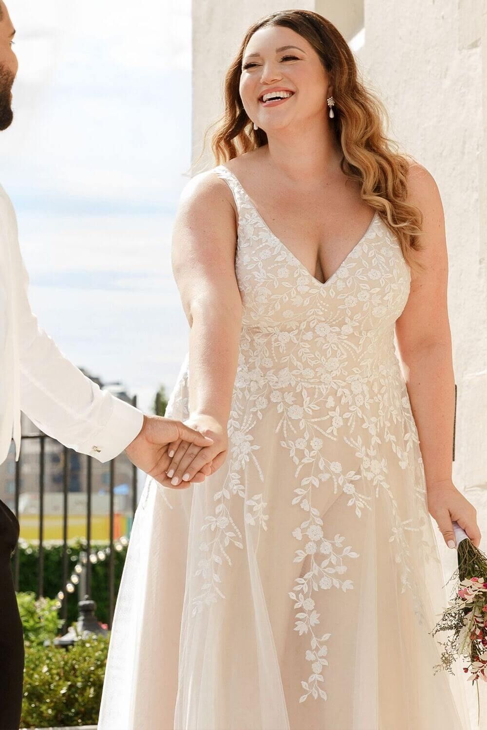 Bride in white dress smiling, holding hands with groom, outdoor setting.