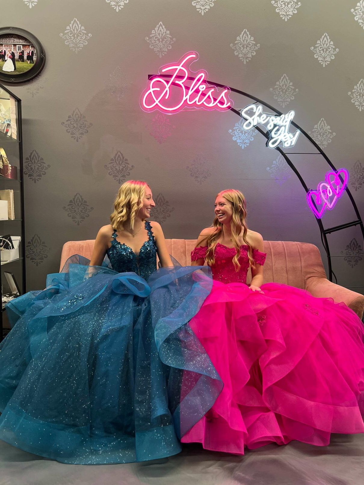 Two women in formal gowns smiling, seated on a pink couch under neon 