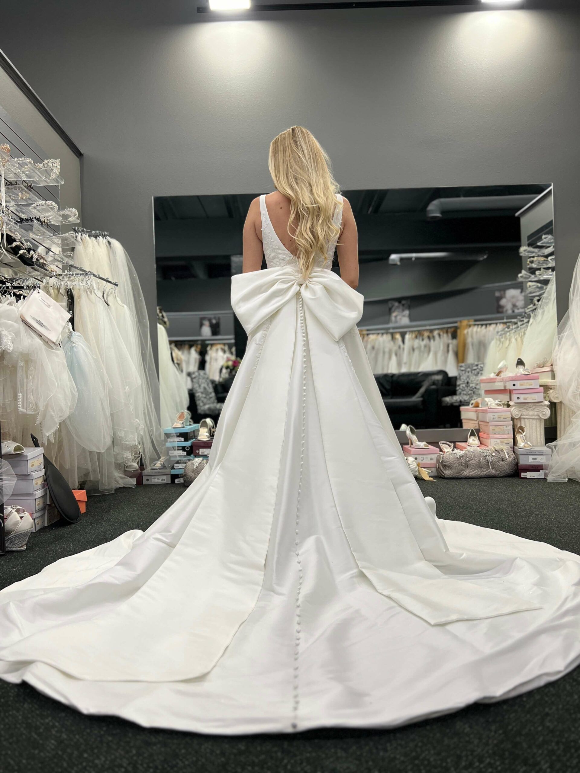 Blonde woman in a white wedding gown with a large bow, standing in a bridal shop.