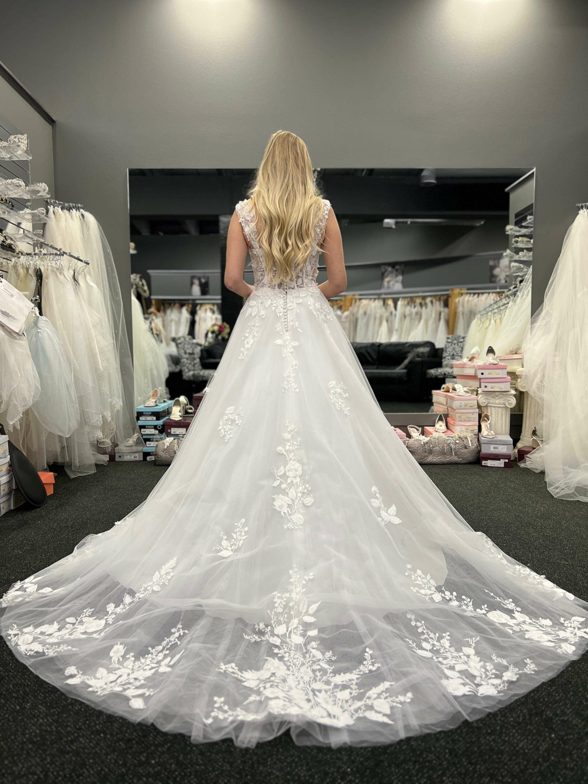 Blonde woman in a white wedding dress with a long train, standing in a bridal shop.