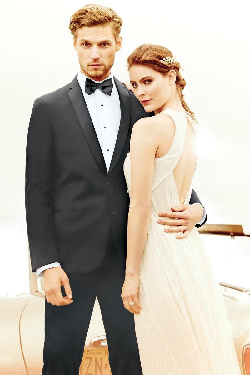 Man in tuxedo with woman in gown, posing near vintage car.