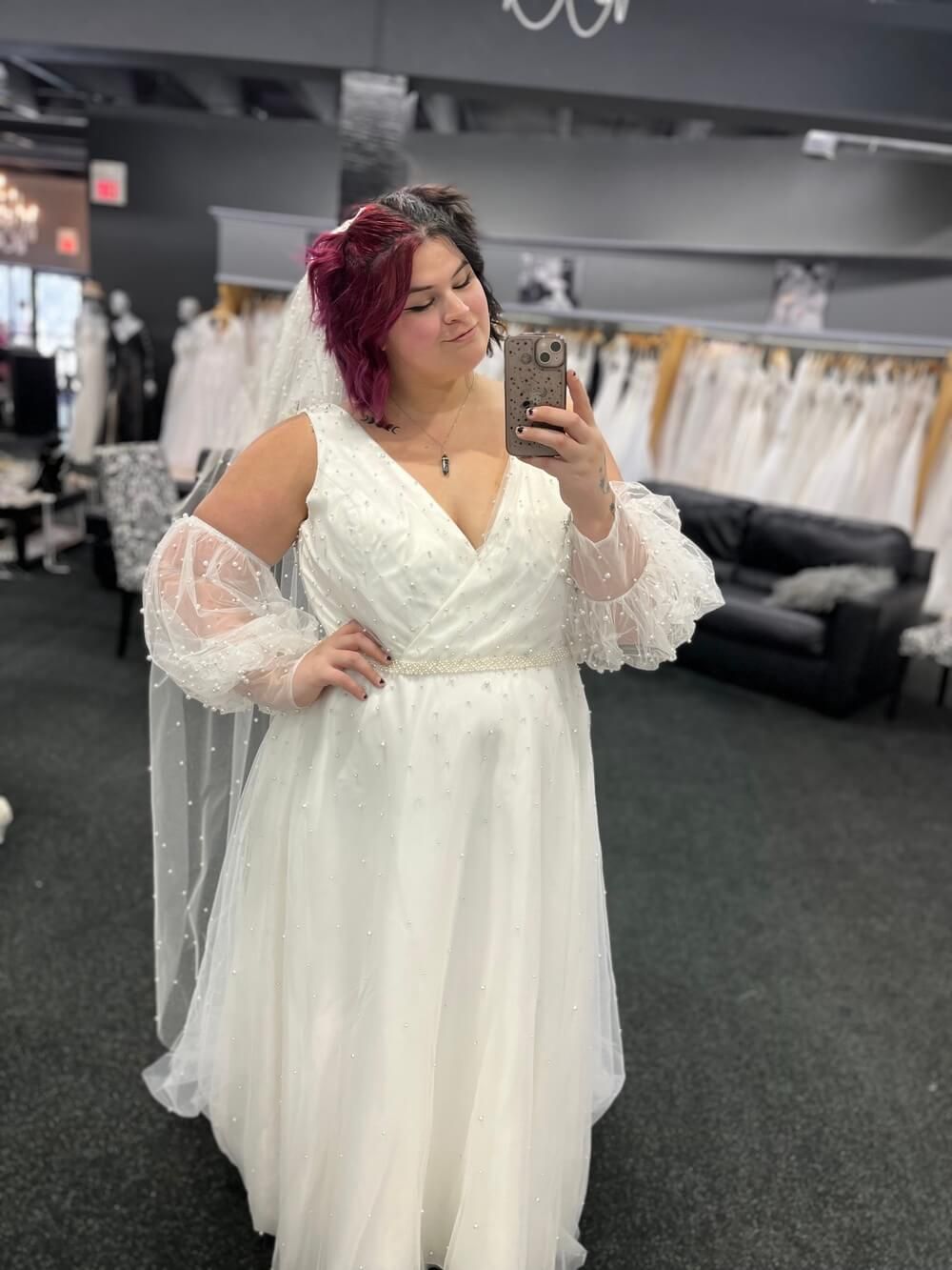 Woman in a white wedding dress with lace sleeves and a veil takes a mirror selfie in a bridal shop.