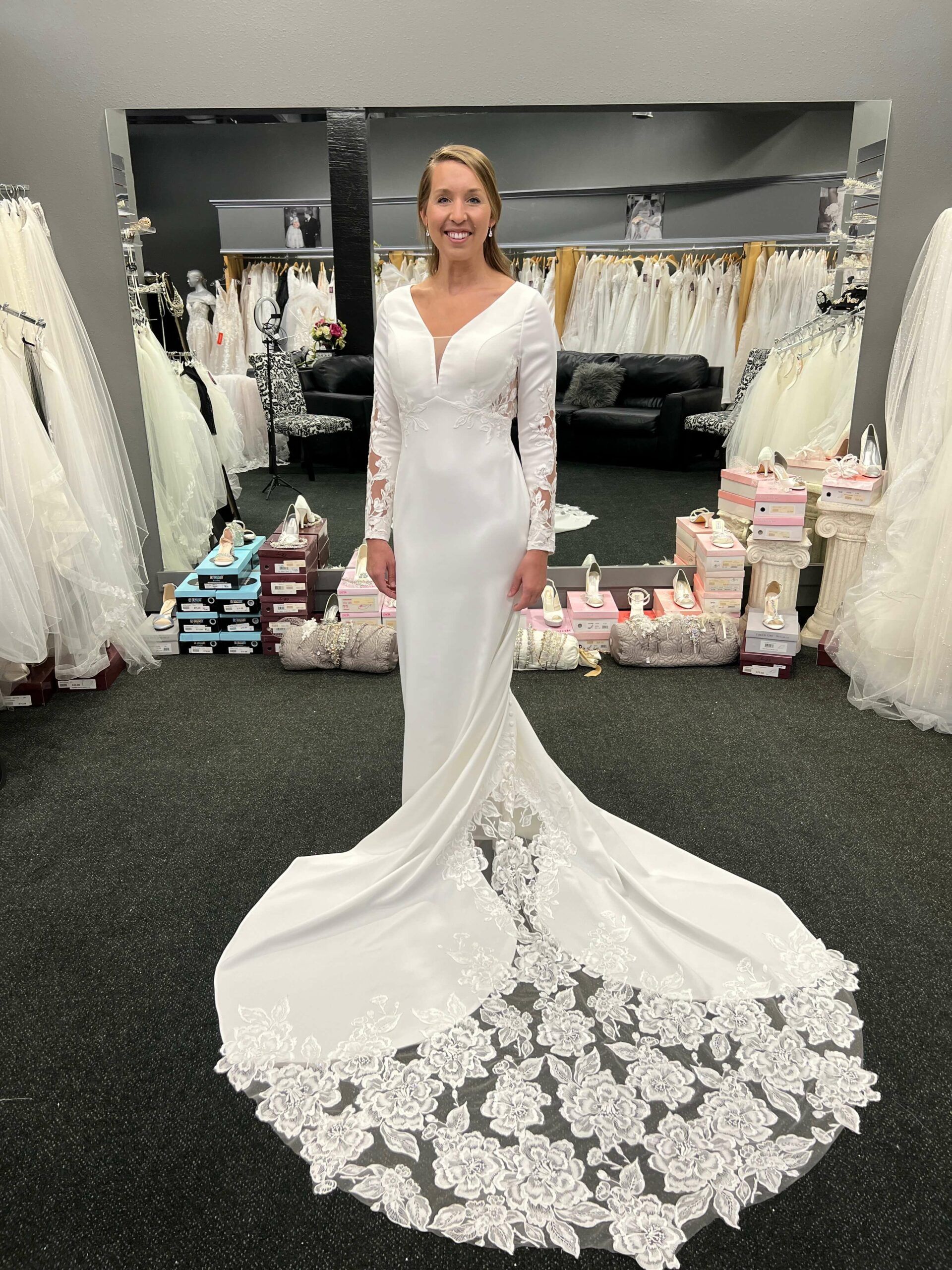 Woman in white wedding dress with long train, standing in a bridal shop.