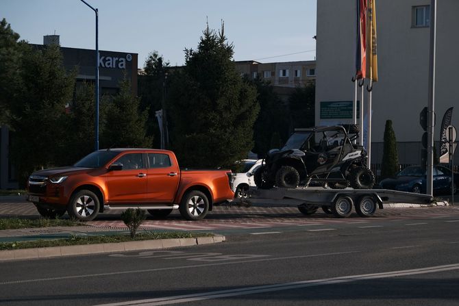 Orange pickup truck towing motorcycles on a trailer beside a road, with buildings in the background