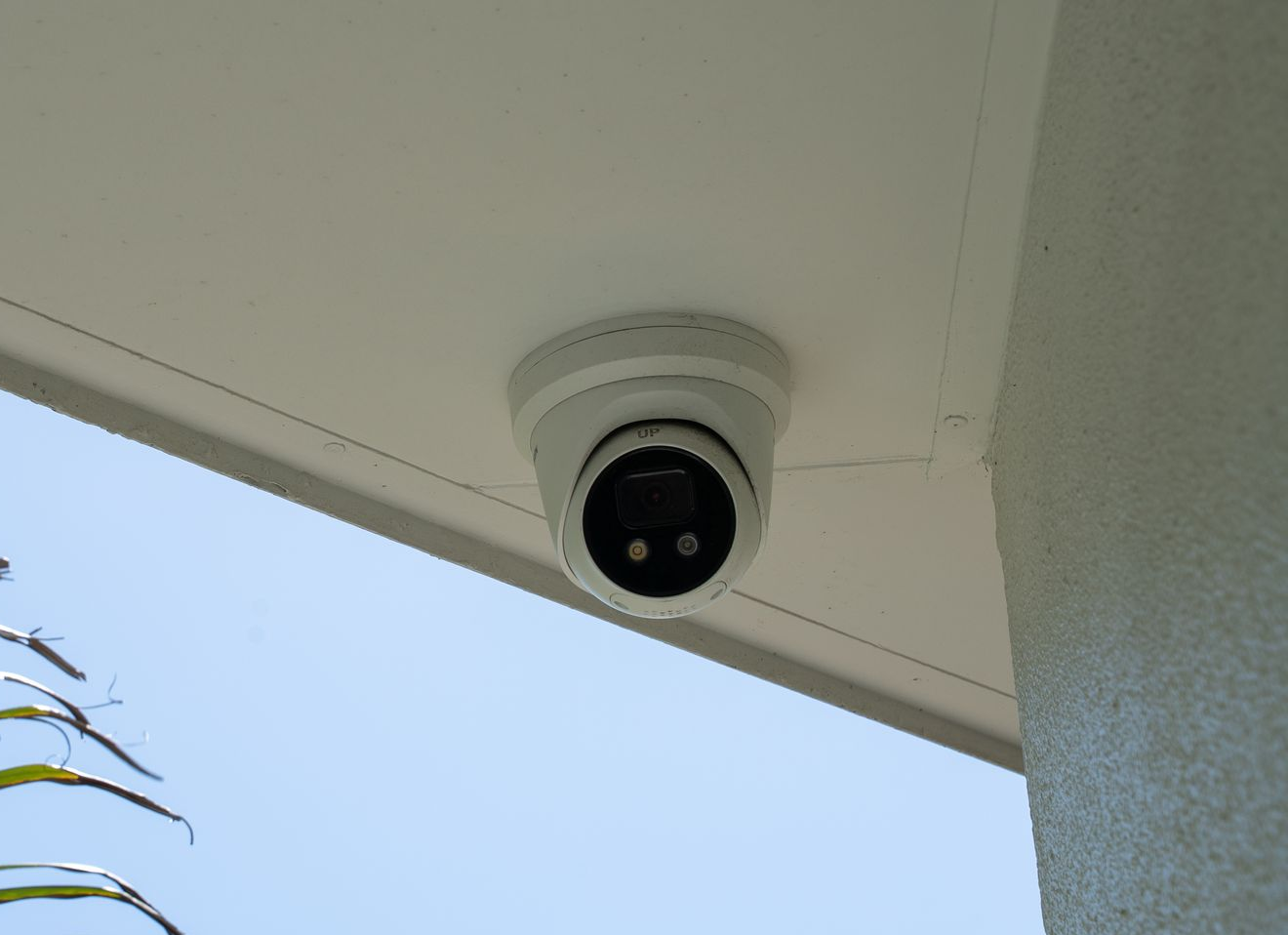 Security Camera Mounted Under a Roof Eave, with Palm Leaves and Blue Sky Nearby — Ralph Ottmann Electrical in Pomona, QLD