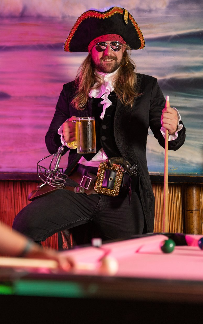 A man dressed as a pirate is standing next to a pool table holding a mug of beer.
