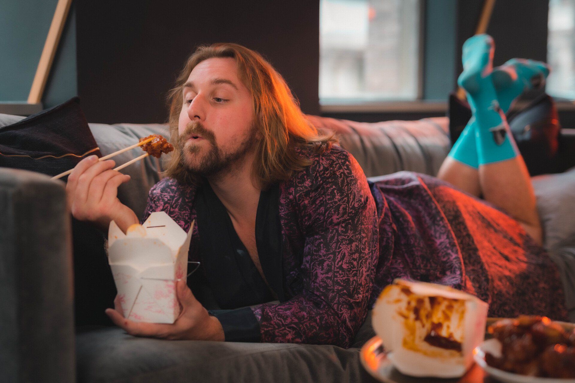 A man is laying on a couch eating food with chopsticks.