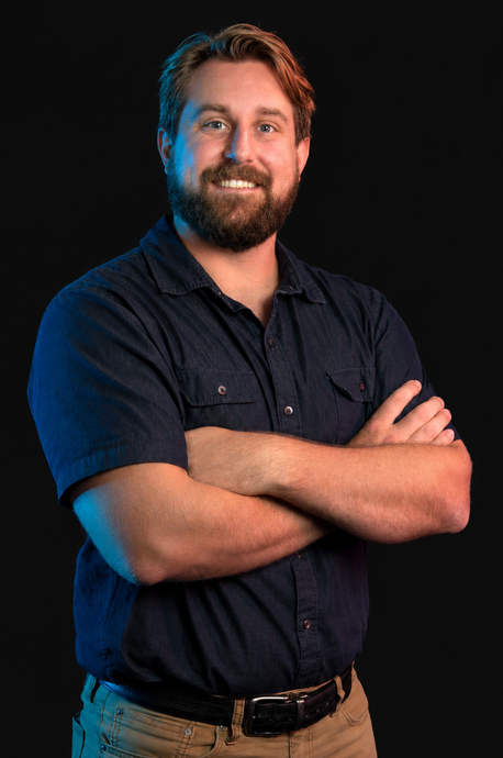 Smiling man with a beard in a navy shirt, arms crossed against a dark background.