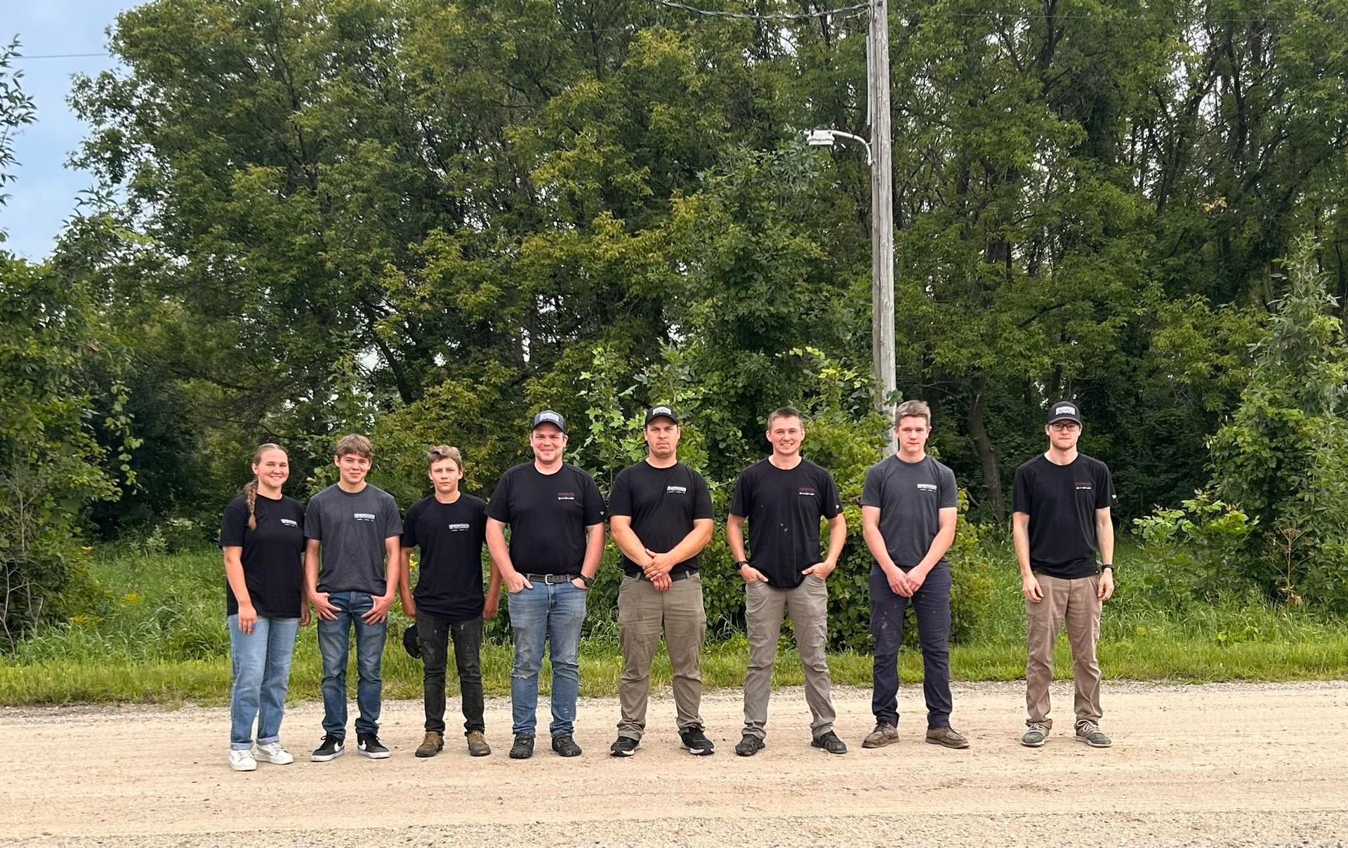 Seven people standing outdoors on a gravel area in front of trees, wearing black shirts and jeans.