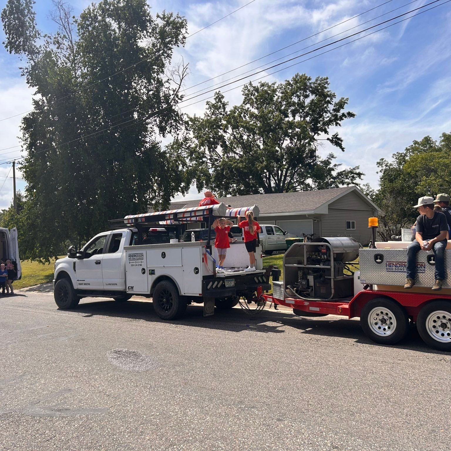 White utility truck with people in red shirts towing a red trailer on a residential street under blue sky