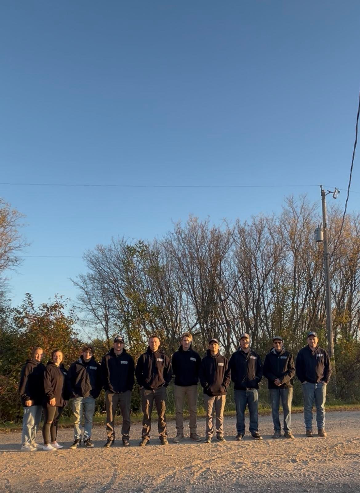 Group of people standing outdoors on a gravel road with trees and a clear blue sky behind them
