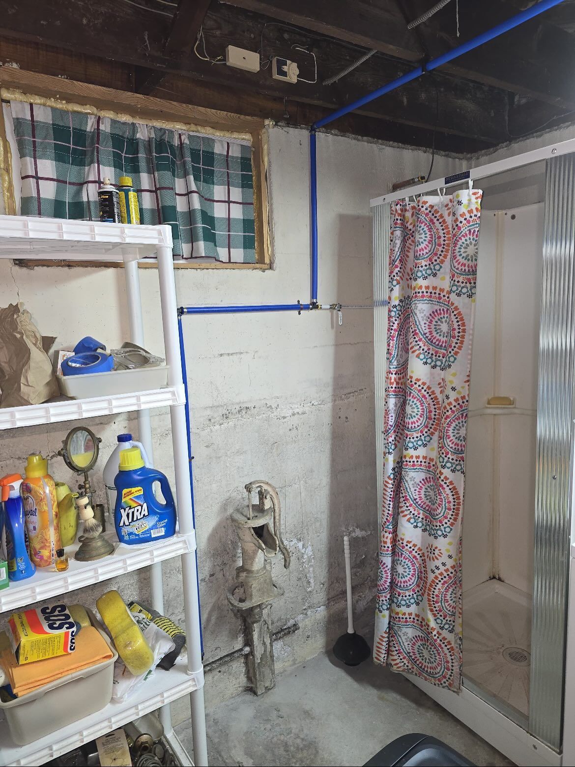 Shelved laundry supplies in a basement utility room with a shower curtain and exposed pipes