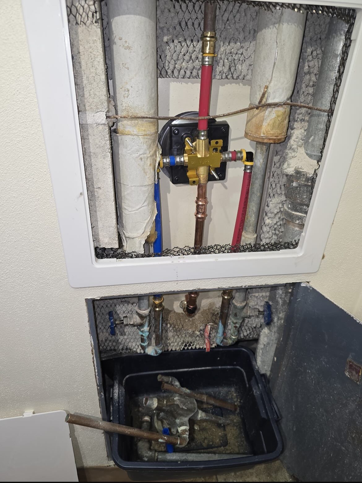 Open plumbing access panel with colored pipes and valves above a floor bucket catching water