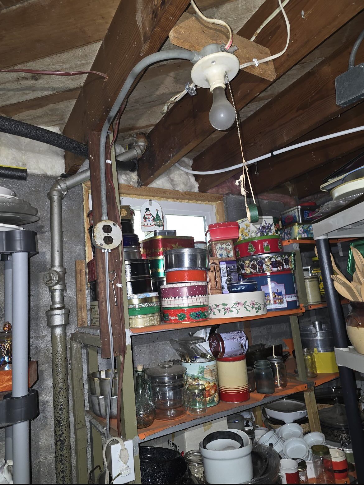 Cluttered basement storage shelf with jars, cans, pipes, and a hanging light bulb under wooden ceiling.