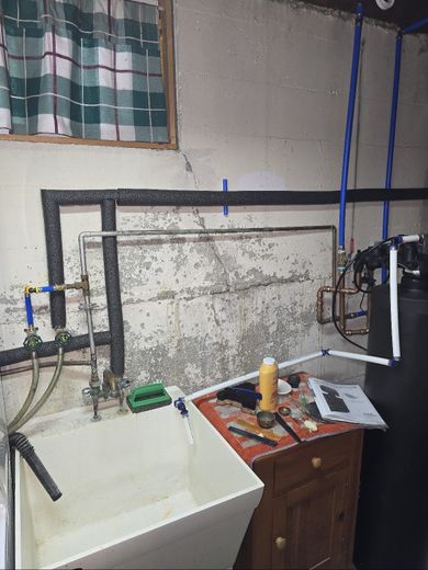 Utility room with sink, exposed pipes, and a cluttered workbench against a stained wall.