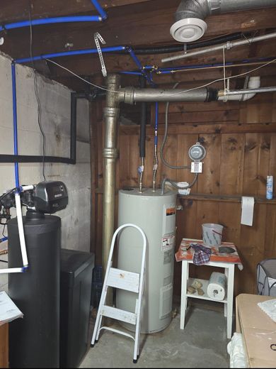 Basement utility room with water heater, exposed pipes, fan, ladder, and wooden wall.