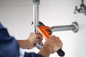 Plumber tightening a metal pipe under a sink with an orange wrench
