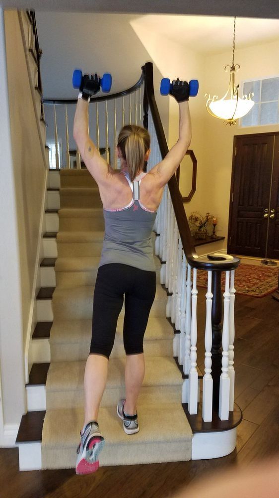 A woman is lifting dumbbells over her head while standing on a set of stairs.