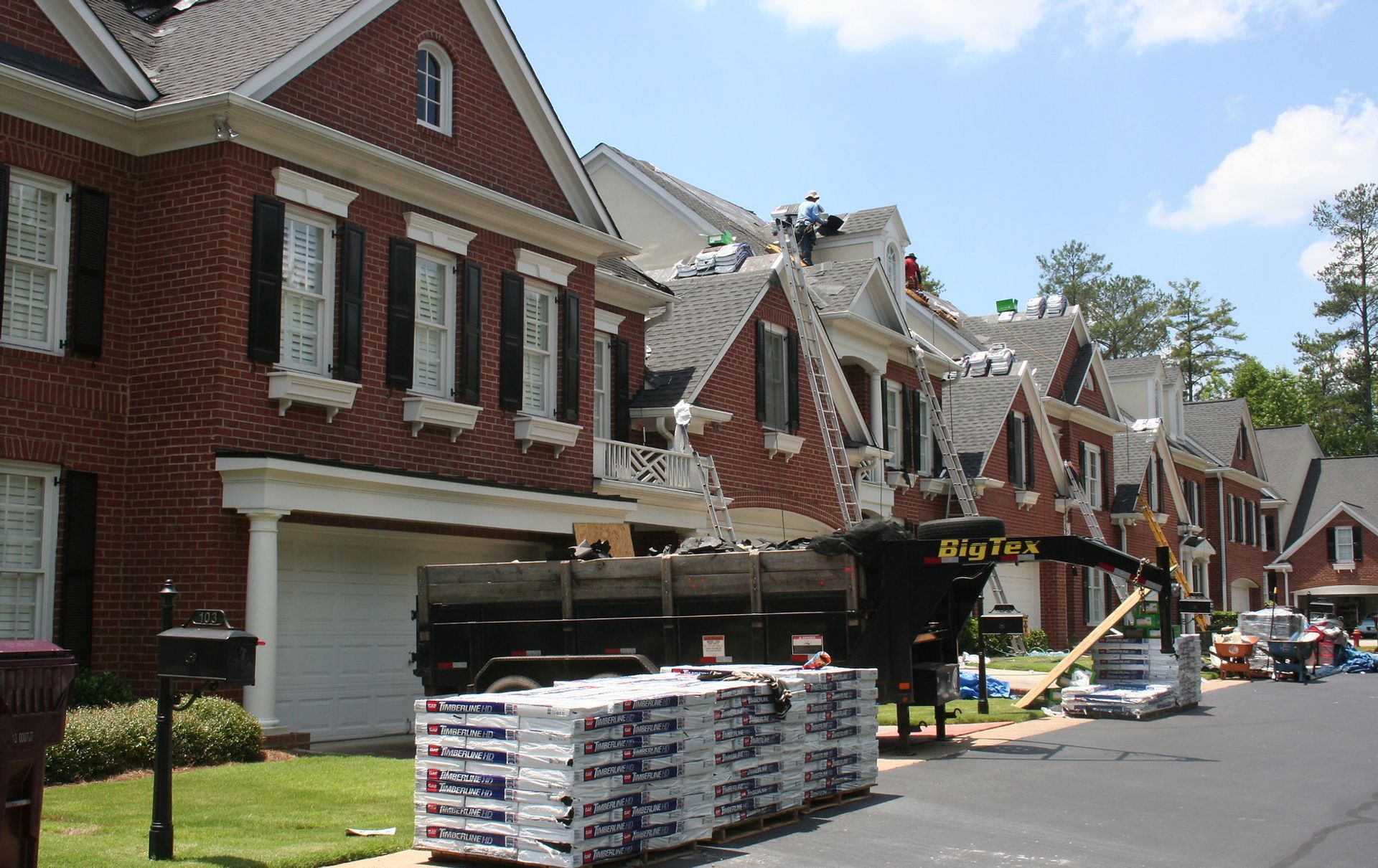 Damaged brick rowhouses with roof repairs, stacked shingles, and workers on a sunny residential street