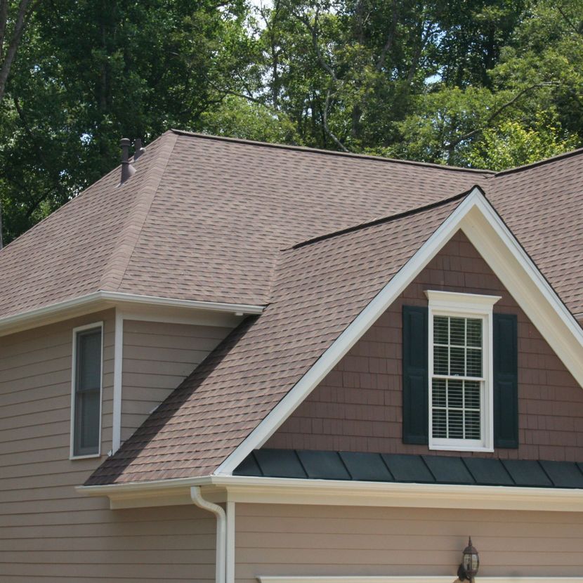 Tan house with brown shingle roof and dormer window against green trees