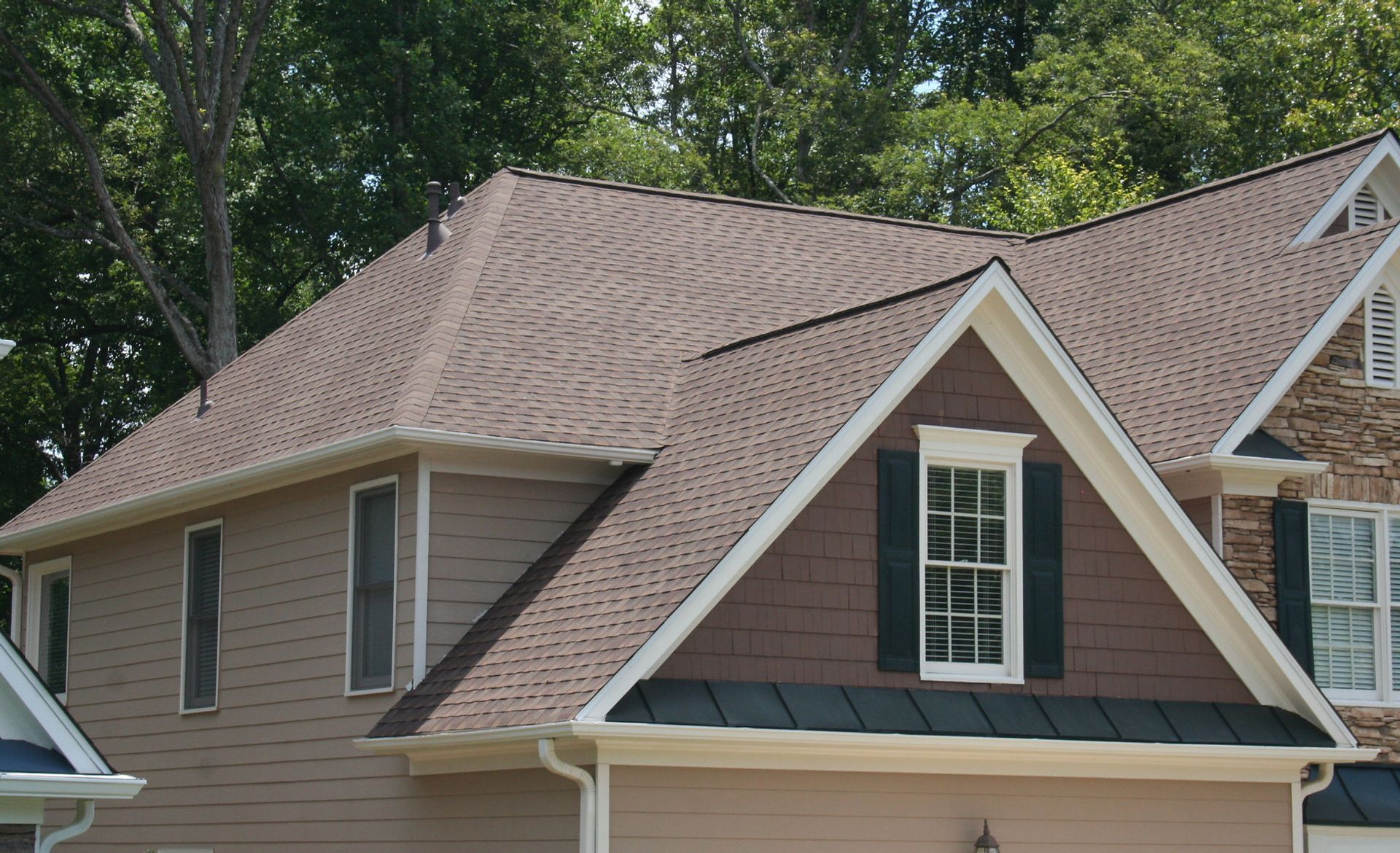 Suburban house with tan siding, brown shingle roof, and a dormer window surrounded by trees.