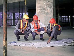Three construction workers in hard hats crouch over blueprints at a building site.