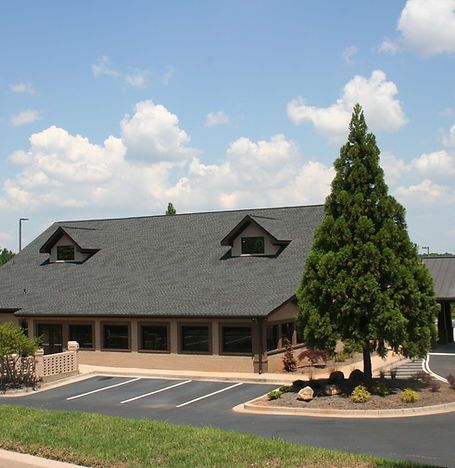 Brick building with dark roof and dormer windows, large tree, and parking lot under a blue sky