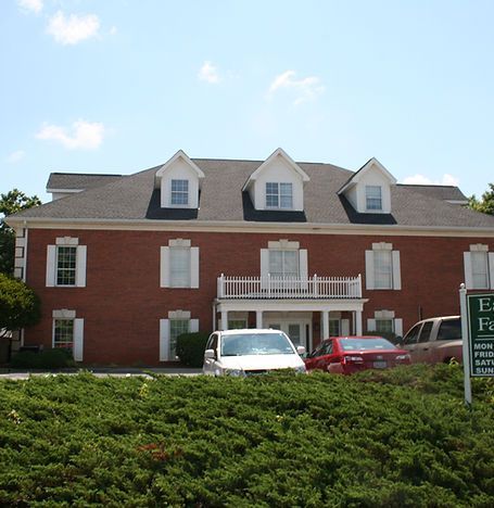 Red brick building with white dormers and a balcony, parked cars in front, and hedge landscaping.