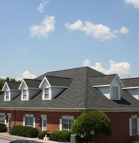 Red brick building with gray shingled roof and dormer windows under a blue sky with clouds