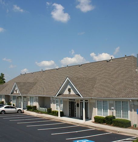 Single-story strip mall with tan roof, white storefronts, and a mostly empty parking lot under a blue sky