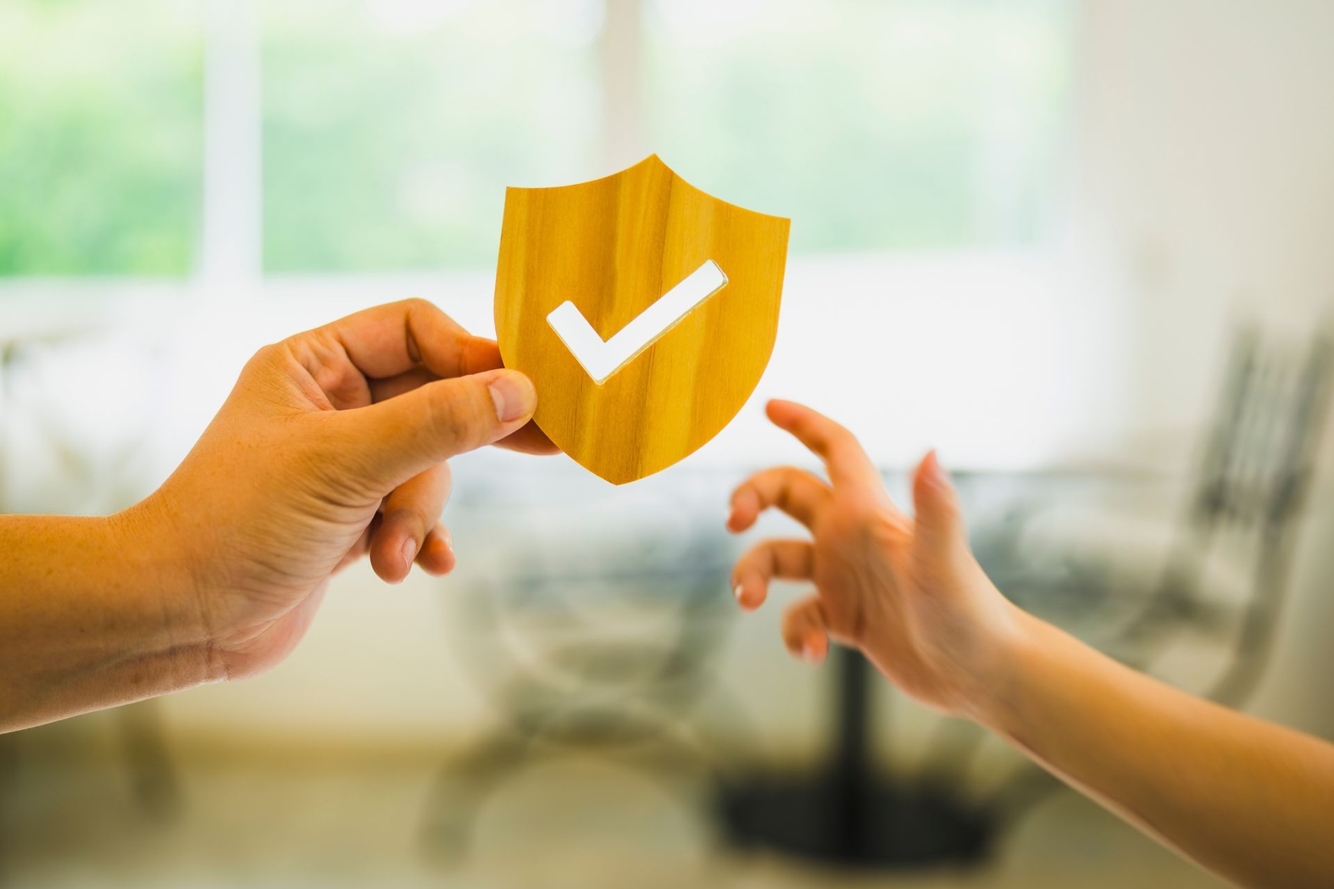 Hands exchanging a gold shield with a white checkmark in a bright blurred room