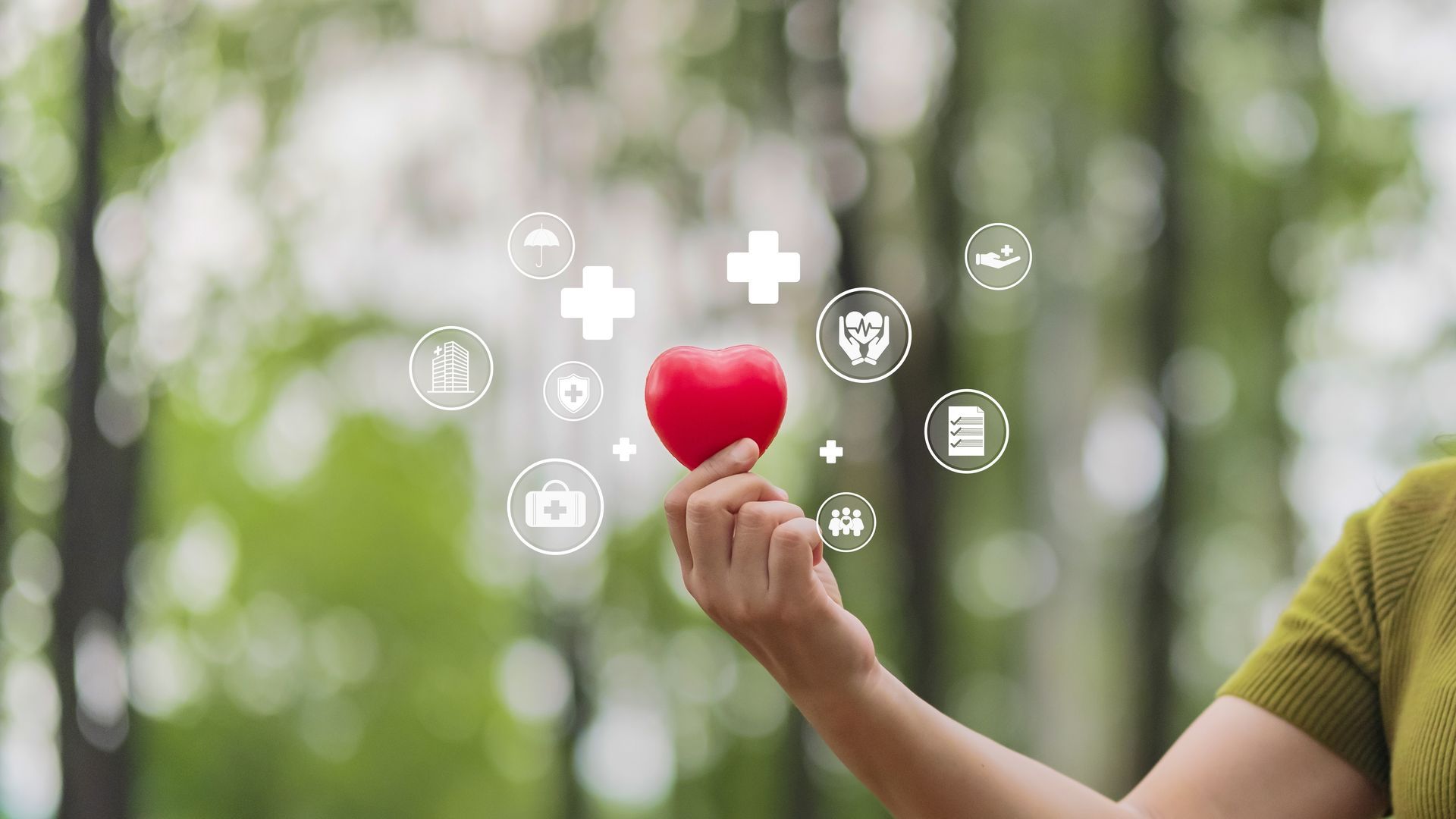 Hand holding a red heart icon with medical symbols floating above against a blurred green background