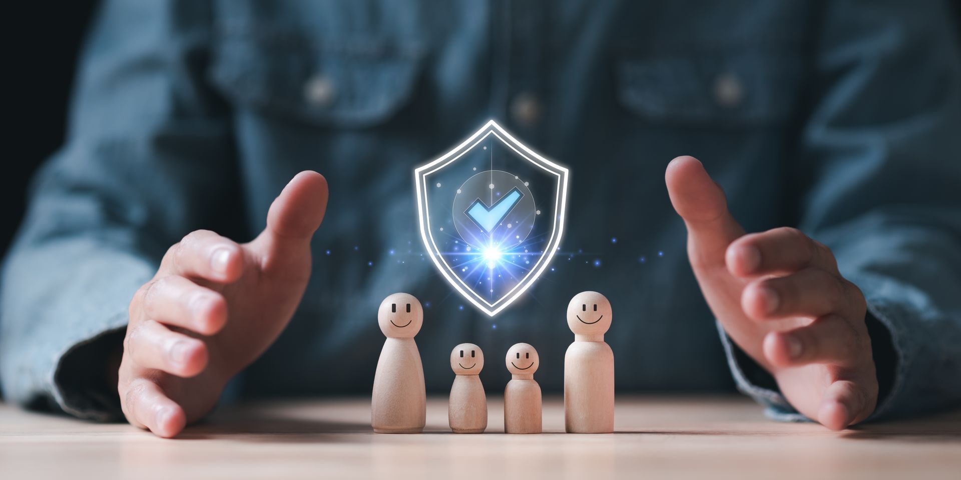 Hands framing a glowing shield above wooden figures on a table, symbolizing protection and security