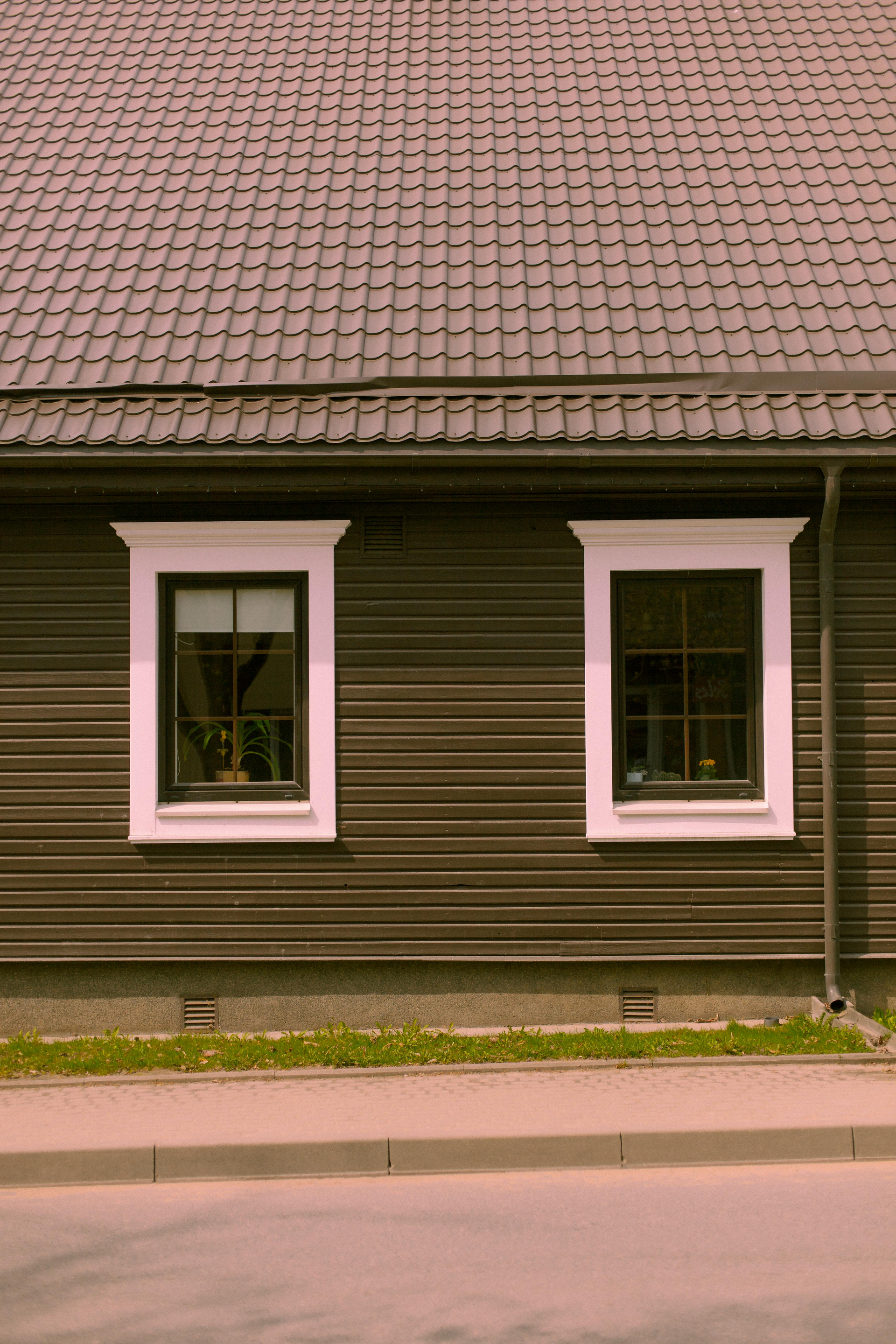 Dark wooden house facade with two white-trimmed windows and a tiled roof beside a street.
