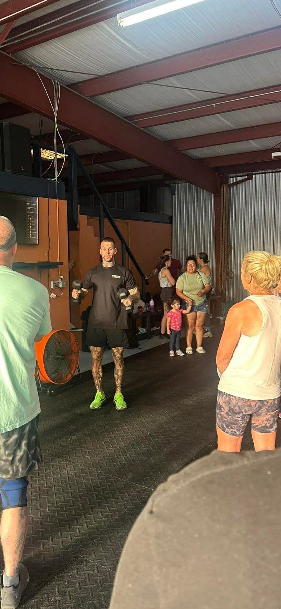 A trainer stands in a gym instructing a small group of people, with a large orange fan visible in the background.