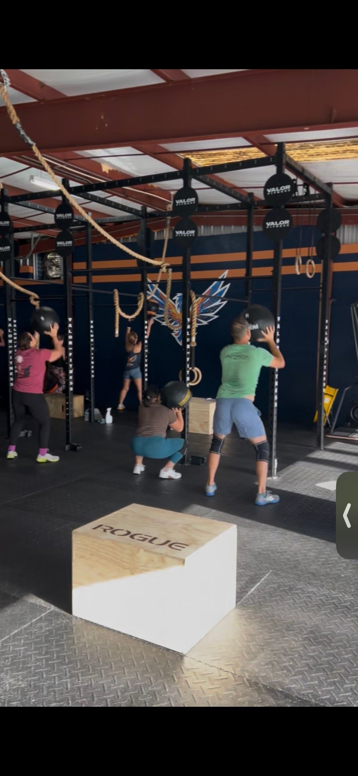 People exercising in a gym, performing wall ball squats with medicine balls in front of a CrossFit rig and plyo boxes.