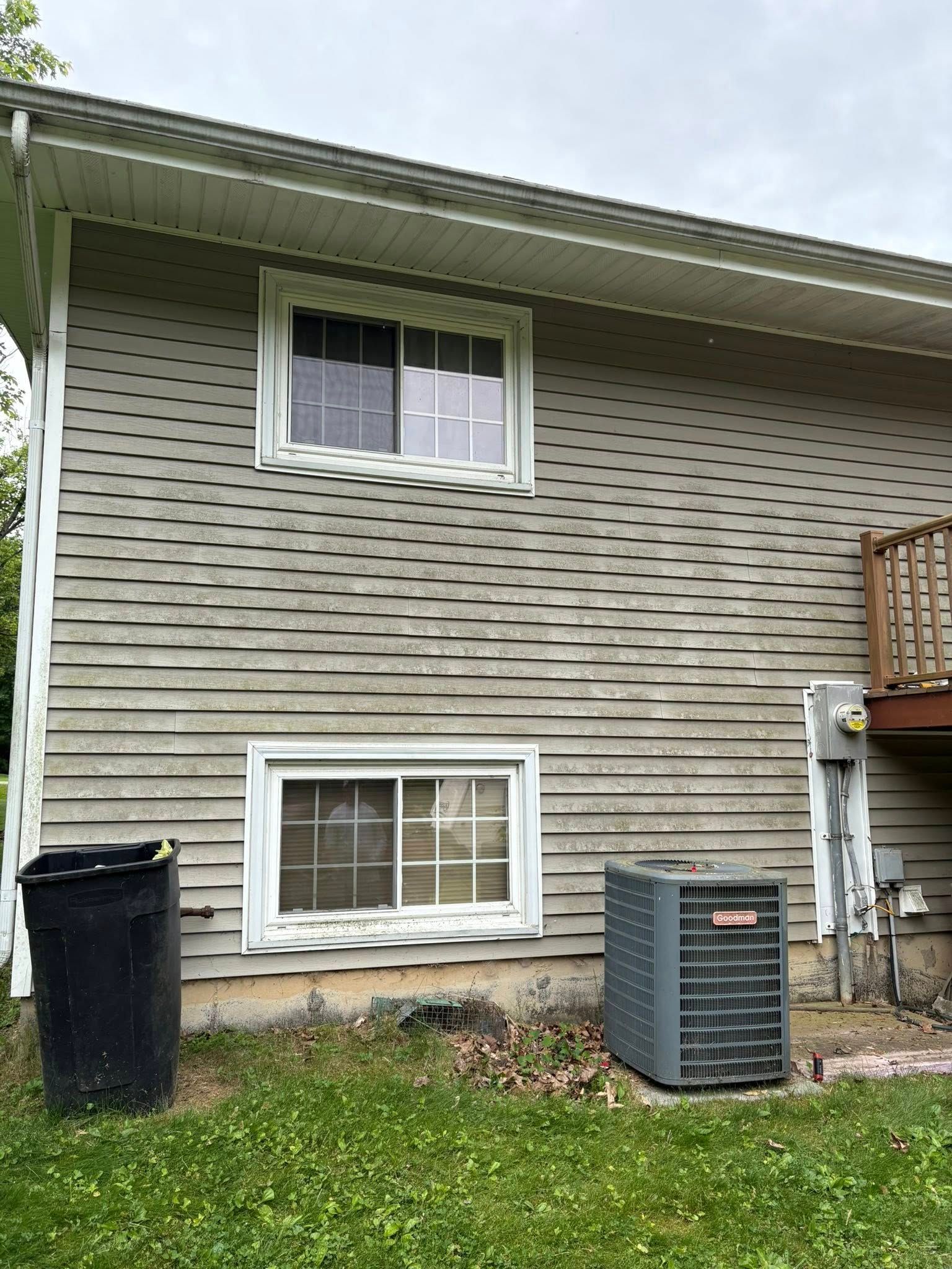 Side of a beige house with two windows, a trash bin, and an air conditioner unit on a grassy yard.