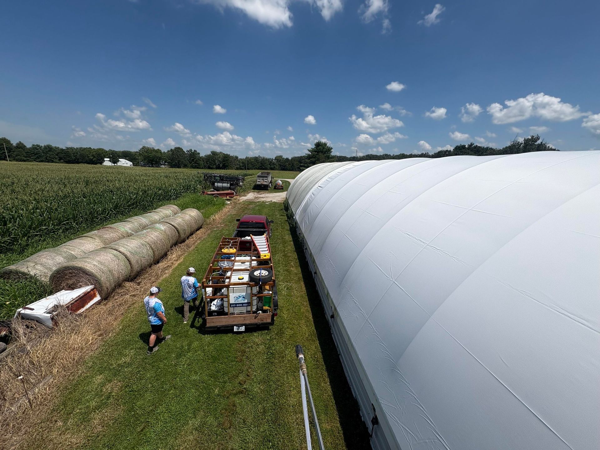 Workers and tractors beside hay bales in a field next to a large white greenhouse under a blue sky