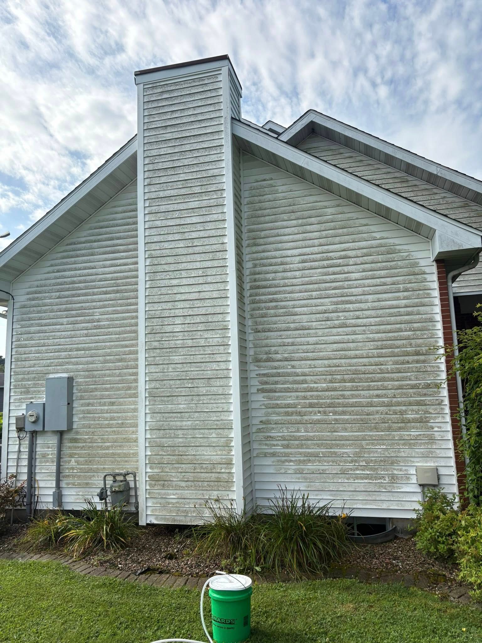Side of a white house with a tall chimney and a green utility meter in front, under a cloudy sky