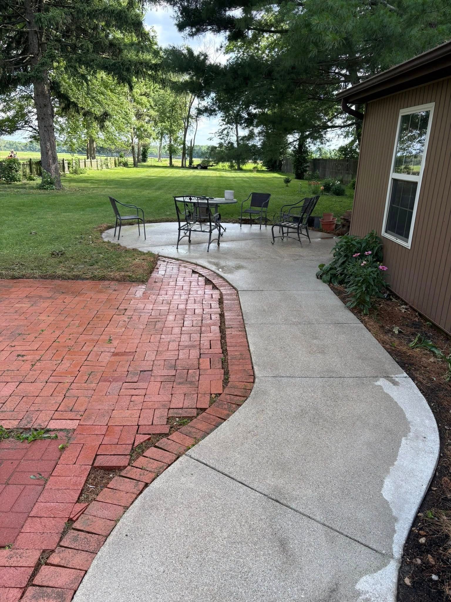Curved concrete walkway beside red brick patio, leading to patio table and chairs under trees by a house.