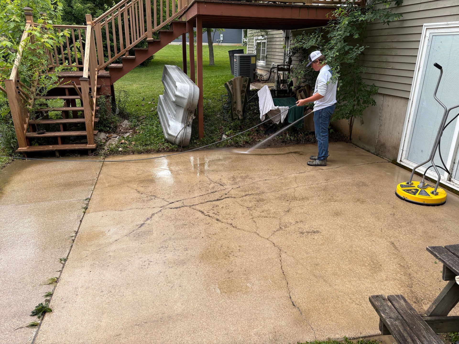 Person pressure washing a wet concrete patio beside a wooden staircase and house exterior